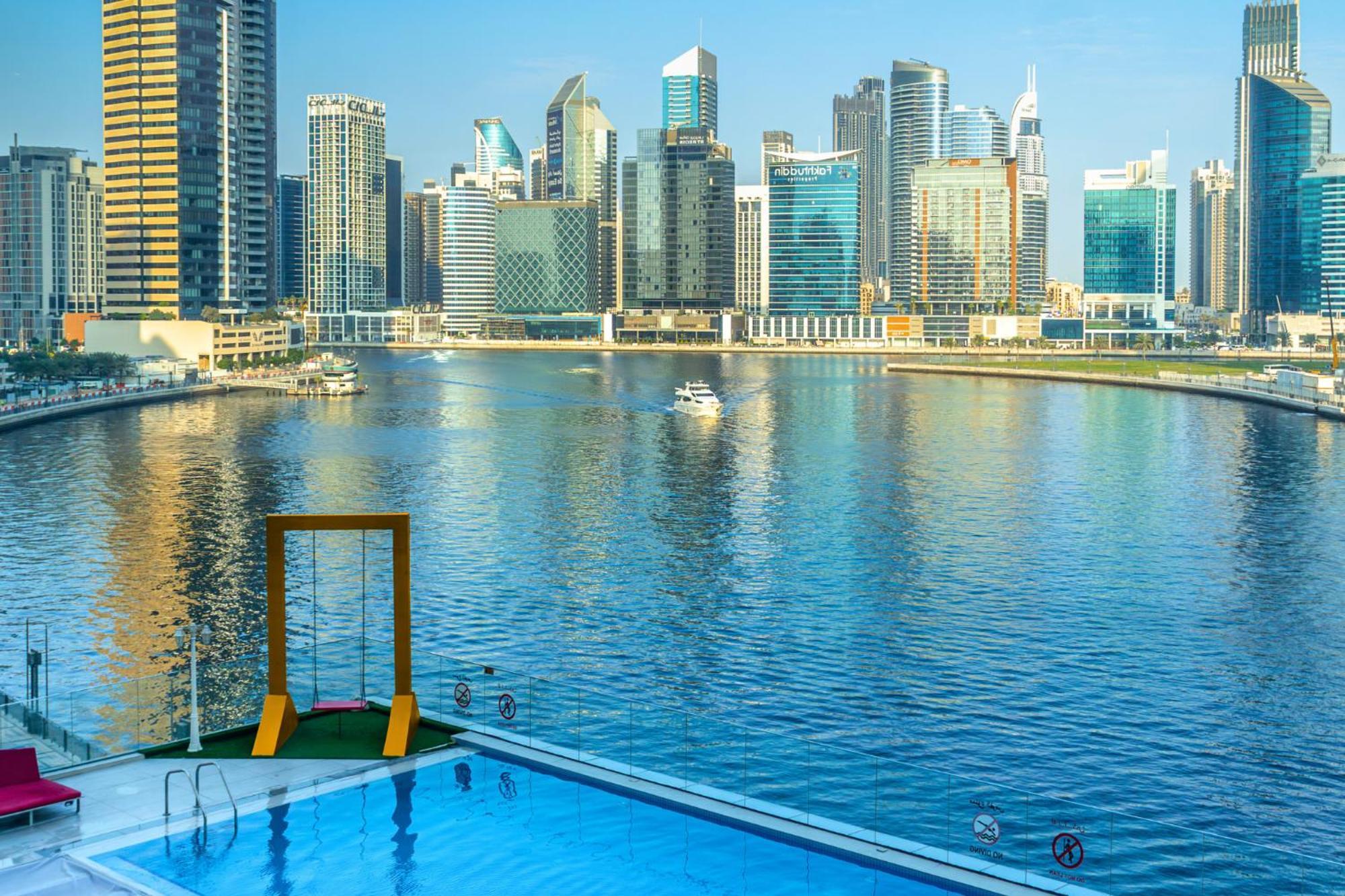 Canal Central Hotel Business Bay Dubai Exterior foto The photo shows a serene waterfront view featuring a modern city skyline with tall buildings reflecting in the water. In the foreground, there is a swimming pool with a swing set beside it, overlooking the calm water. The skyline is characterized by 