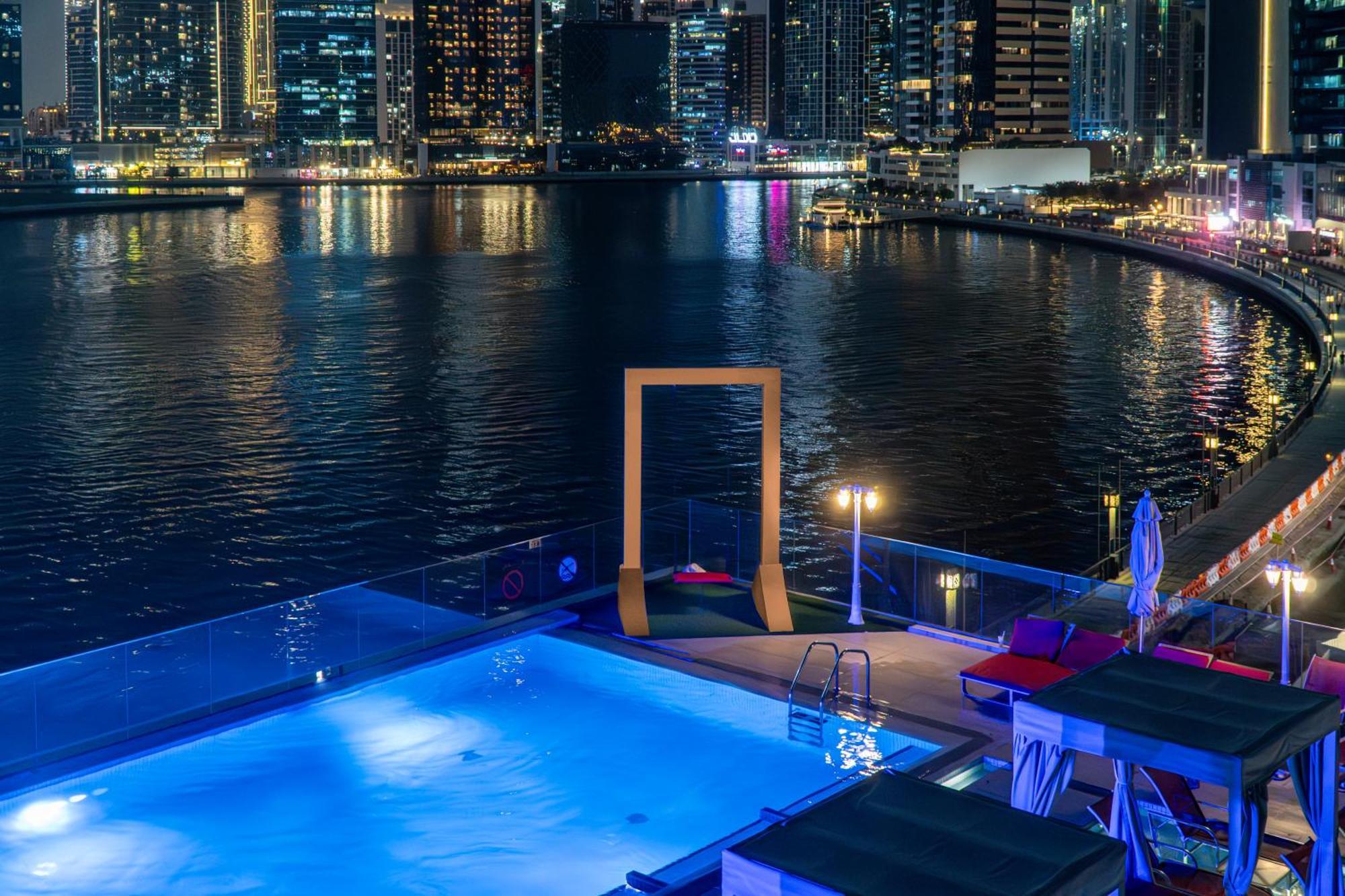 Canal Central Hotel Business Bay Dubai Exterior foto The photo shows a nighttime view of a modern pool area overlooking a body of water, likely a marina or waterfront. The pool is illuminated with a blue light, and there are lounge chairs beside it. In the background, a city skyline can be seen with ta