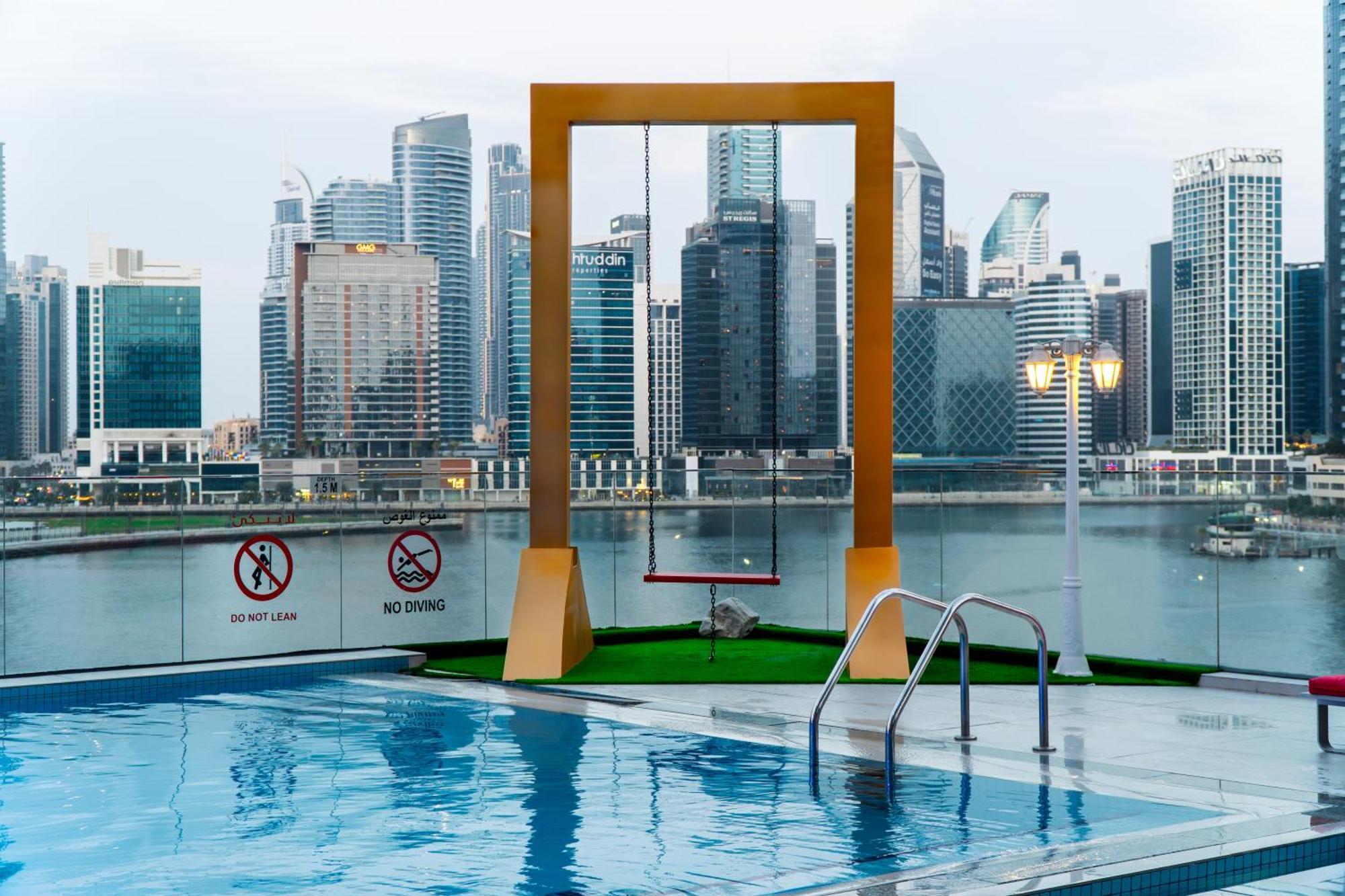 Canal Central Hotel Business Bay Dubai Exterior foto The photo depicts a modern rooftop pool area featuring a clear blue swimming pool with a ladder. In the background, there are tall city buildings, indicating a vibrant urban setting. Prominently, a large, yellow swing set is positioned next to the po
