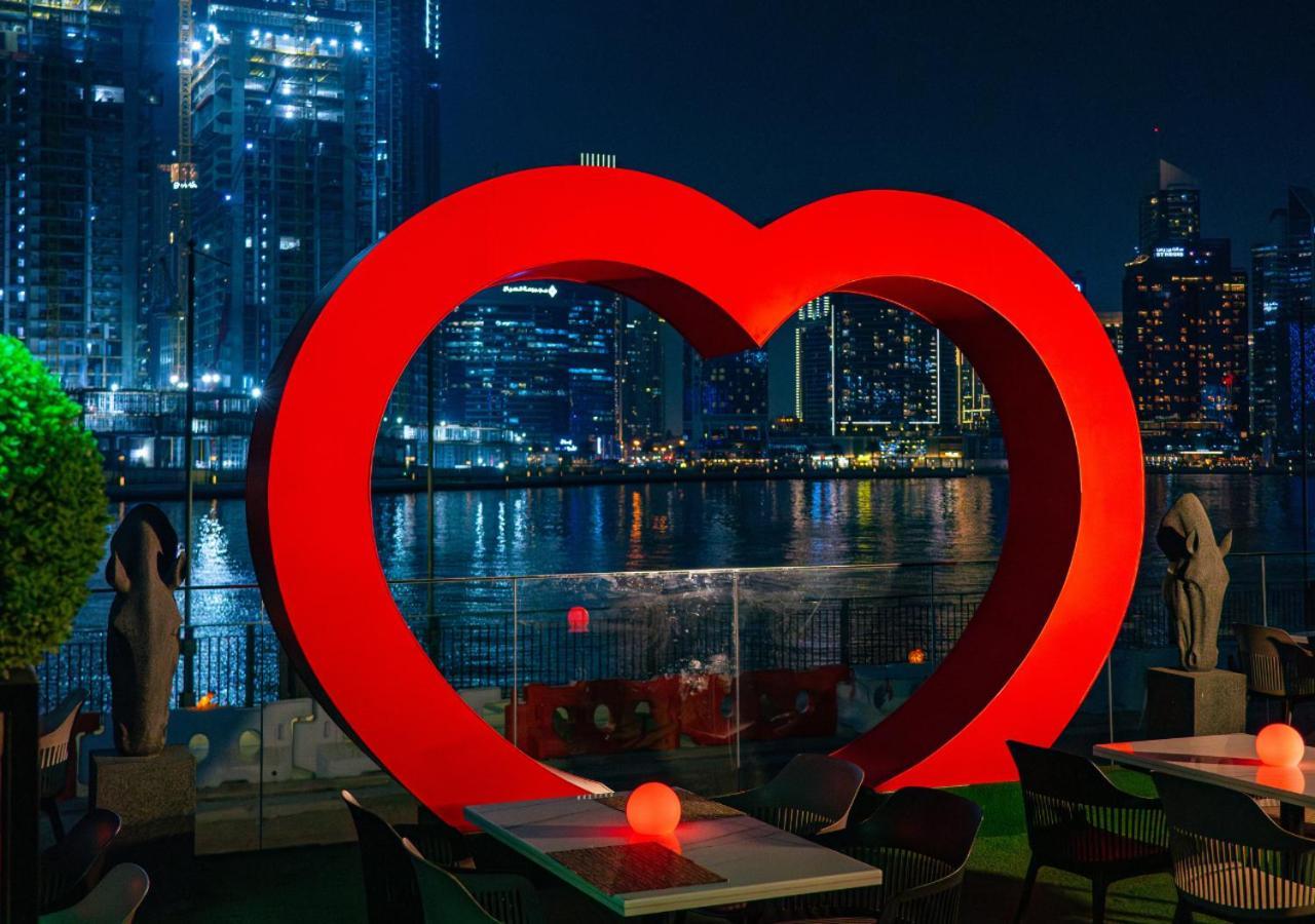 Canal Central Hotel Business Bay Dubai Exterior foto The photo depicts a large, illuminated red heart sculpture set against a nighttime urban backdrop. The heart is prominently displayed near a body of water, with city skyline buildings and lights reflecting off the surface. There are tables in the for