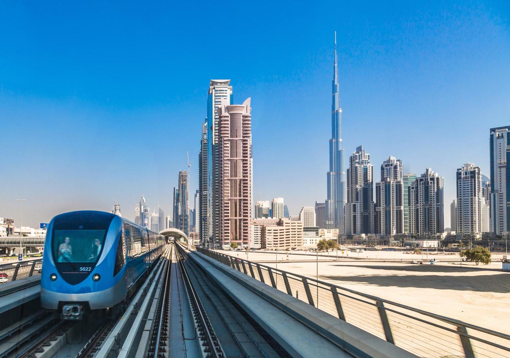 Canal Central Hotel Business Bay Dubai Exterior foto The photo shows a modern metro train on elevated tracks, traveling through a cityscape. To the right, there's a cluster of tall skyscrapers, one of which is recognizable as the Burj Khalifa, the tallest building in the world. The sky is clear and blu